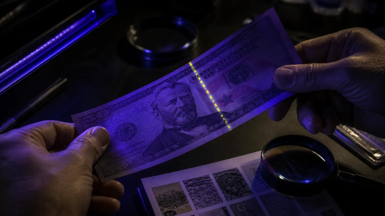A person examining a $50 bill with a UV light, highlighting security features.